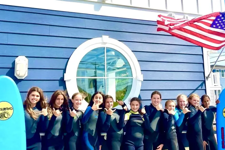 Group of people in wetsuits holding surfboards in front of LBI Surfing building with a US flag.