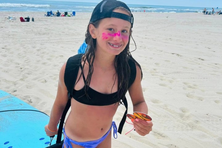 Girl on beach with a surfboard, wearing a cap and pink face paint, under a sunny sky.