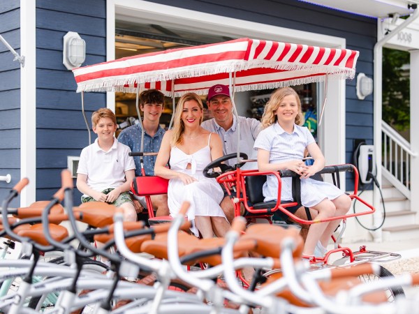 Family of five sitting on a red four-wheel bike with striped canopy, smiling outdoors with bikes in foreground.