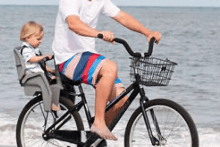 Man biking on beach with child in rear seat and wearing a cap.